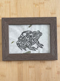 Framed artwork of a frog with a hiking bag on its back on a wooden surface