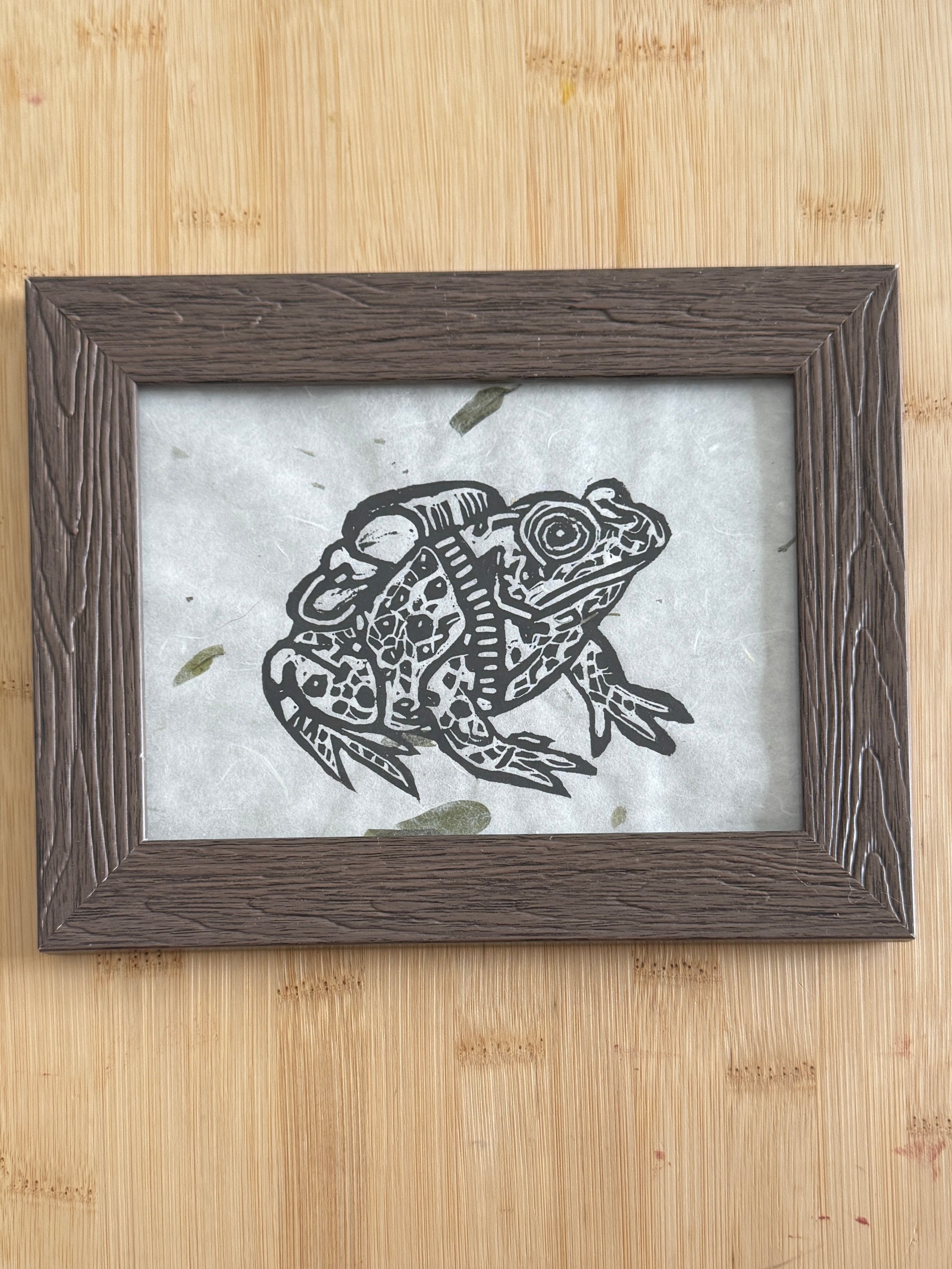 Framed artwork of a frog with a hiking bag on its back on a wooden surface