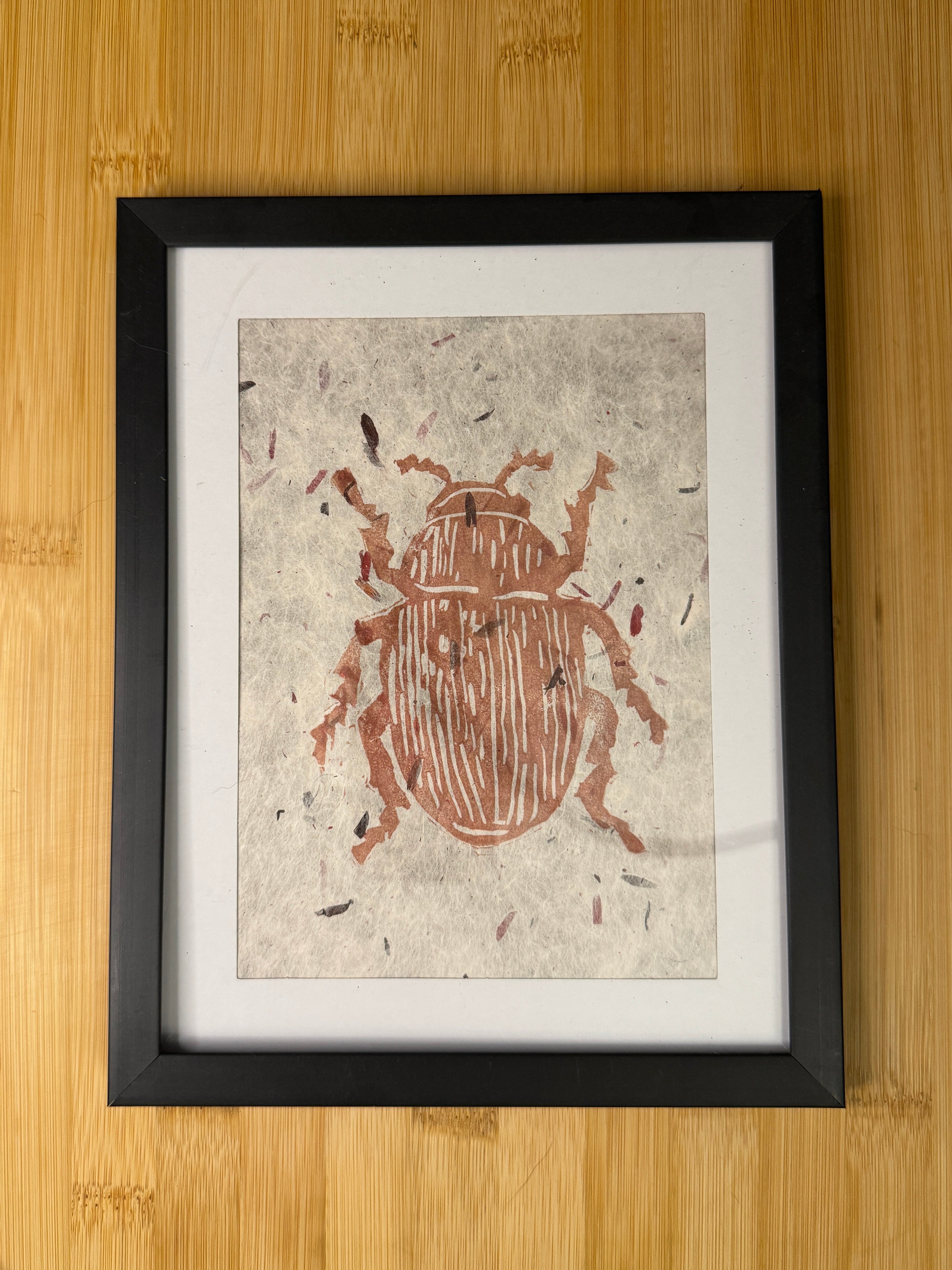 Framed artwork of a beetle on textured paper with a black frame, placed on a wooden surface.