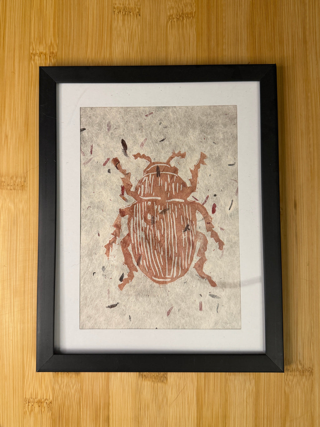 Framed artwork of a beetle on textured paper with a black frame, placed on a wooden surface.