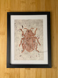 Framed artwork of a beetle on textured paper with a black frame, placed on a wooden surface.