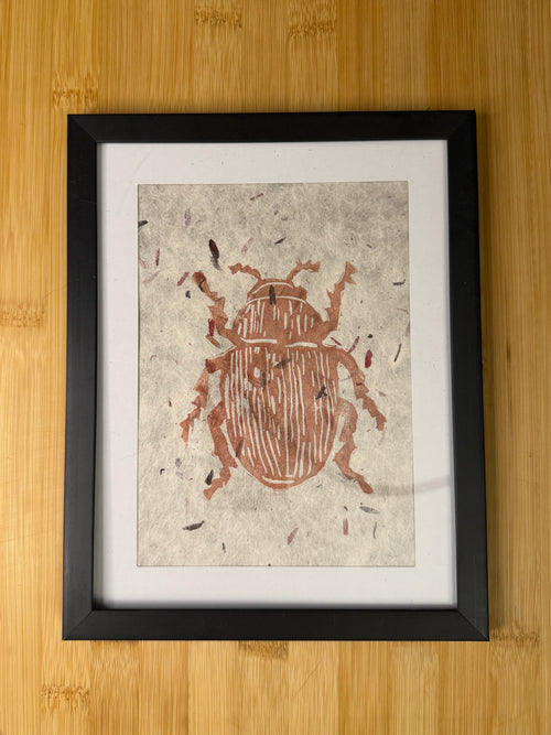 Framed artwork of a beetle on textured paper with a black frame, placed on a wooden surface.